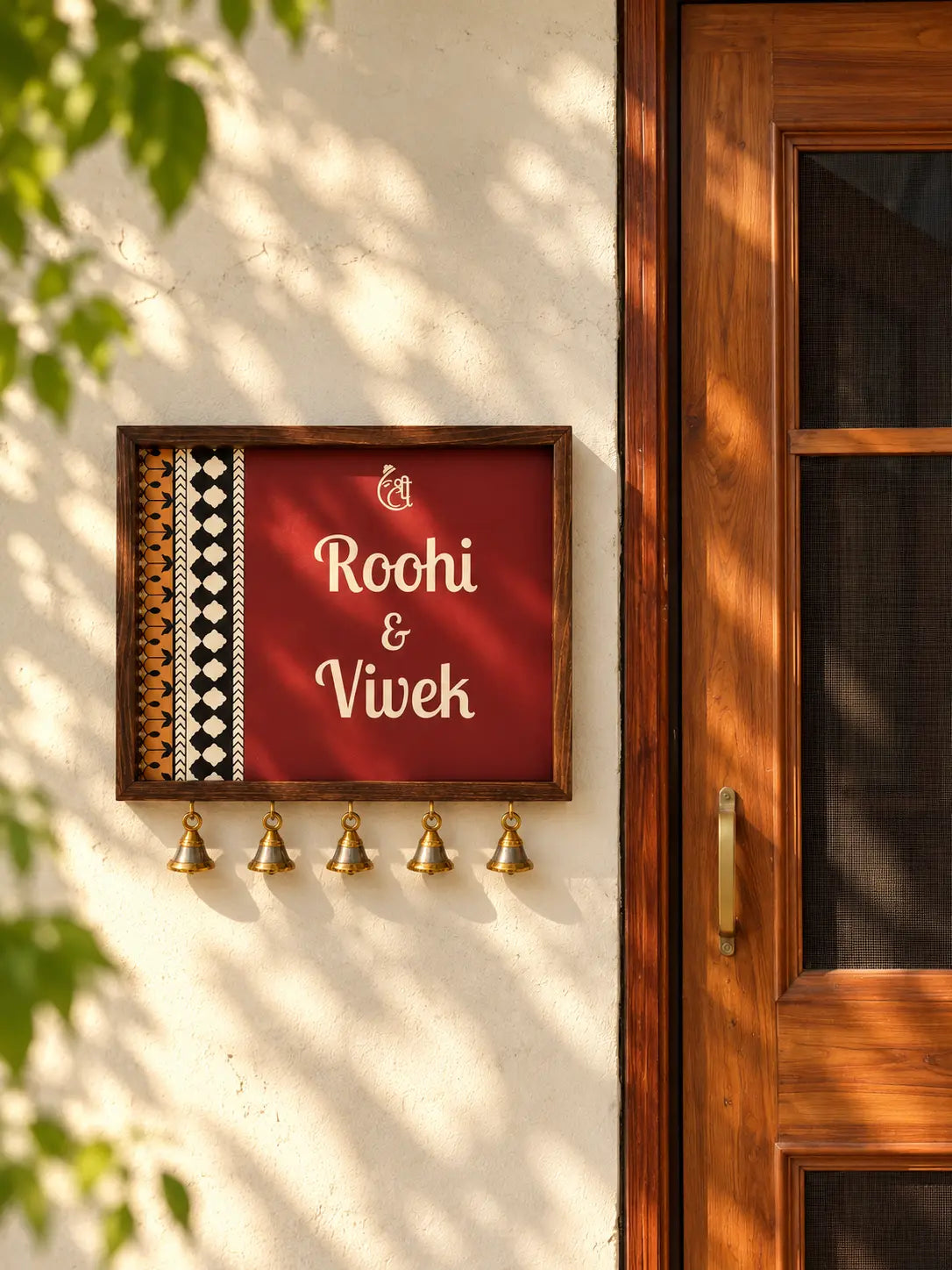 Maroon ethnic name plate with Shree Ganesh motif, wooden frame and brass bells beside a wooden door
