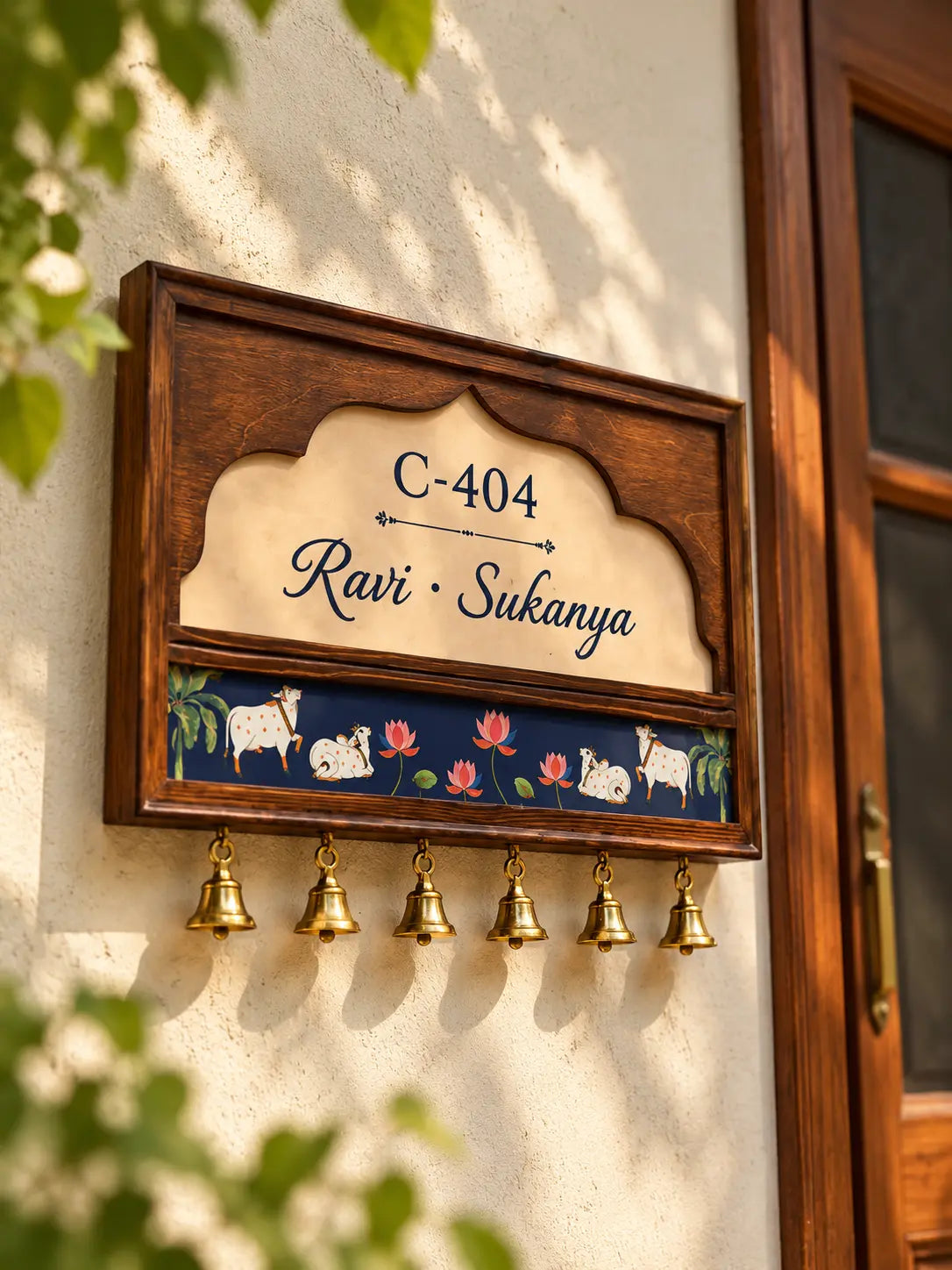 Wooden name plate with cream arch panel, Pichwai cow and lotus band and six brass bells beside a wooden door